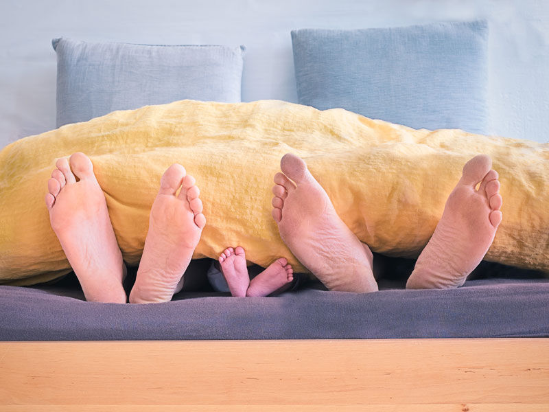 Zeigt die Fußsohlen einer drei-köpfigen Familie, die zugedeckt in einem Doppelbett liegen