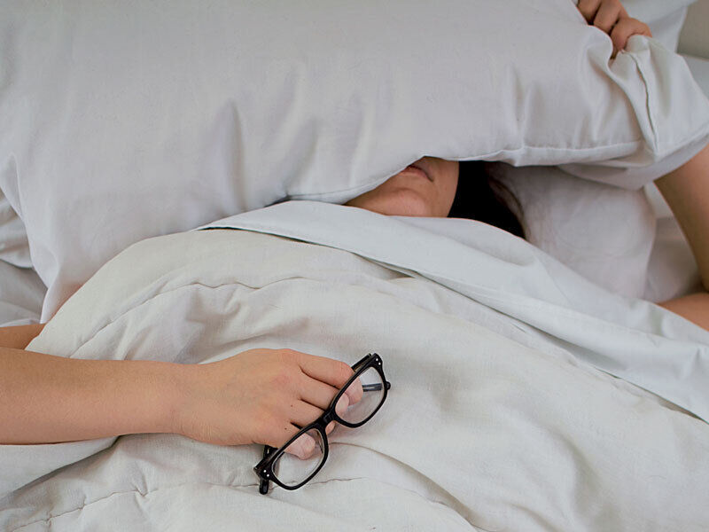 Frau, die im Bett liegt, mit Kissen auf dem Gesicht und Brille in der Hand